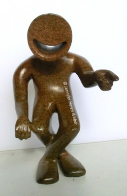 Das Bild zeigt eine braune Specksteinfigur: eine lachende Person, die mit ausgestrecker Hand auf jemanden zeigt.