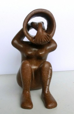 Das Bild zeigt eine braune Specksteinfigur: eine Frau, deren verschränkte Hände von hinten durch den ausgehöhlten Kopf nach vorne gestreckt sind.