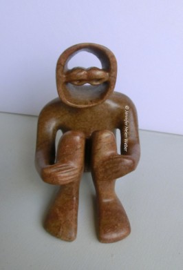 Die Specksteinfigur zeigt eine kauerne Figur, die missmutig dreinschaut.